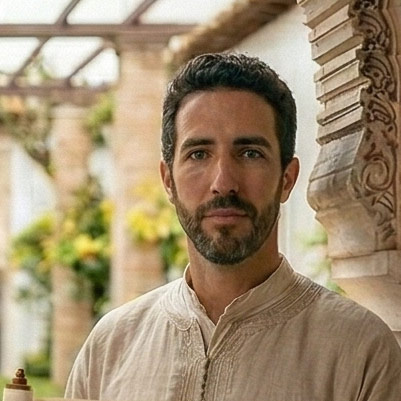 Man with short dark hair and beard wearing a beige traditional-style shirt, standing near an ornate stone column with blurred greenery in the background.