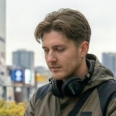 Young man with short brown hair wearing a jacket and black headphones around his neck, looking down with a blurred cityscape background.