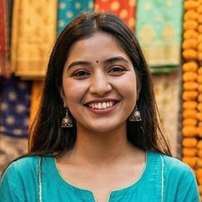 Smiling woman with long dark hair wearing traditional jhumka earrings and a teal outfit standing in front of colorful textile background.
