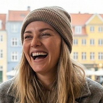 Smiling woman with long blonde hair wearing a brown knit beanie and gray jacket outdoors with buildings in the background.